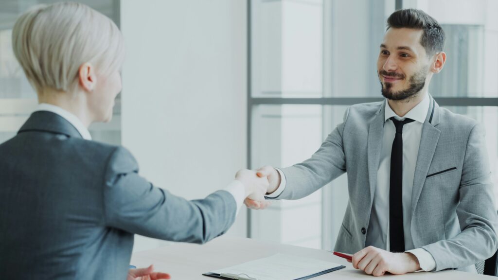Twee professionals hebben contact met elkaar doordat zij elkaar een hand geven over een bureau in een kantooromgeving.