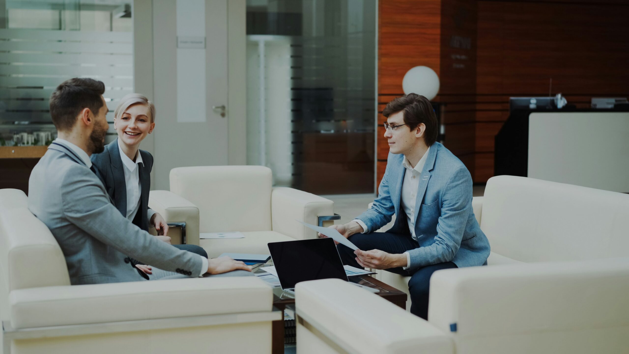Drie professionals in informeel overleg over Governance op witte fauteuils in een moderne kantoorlobby, met een laptop en documenten op tafel.